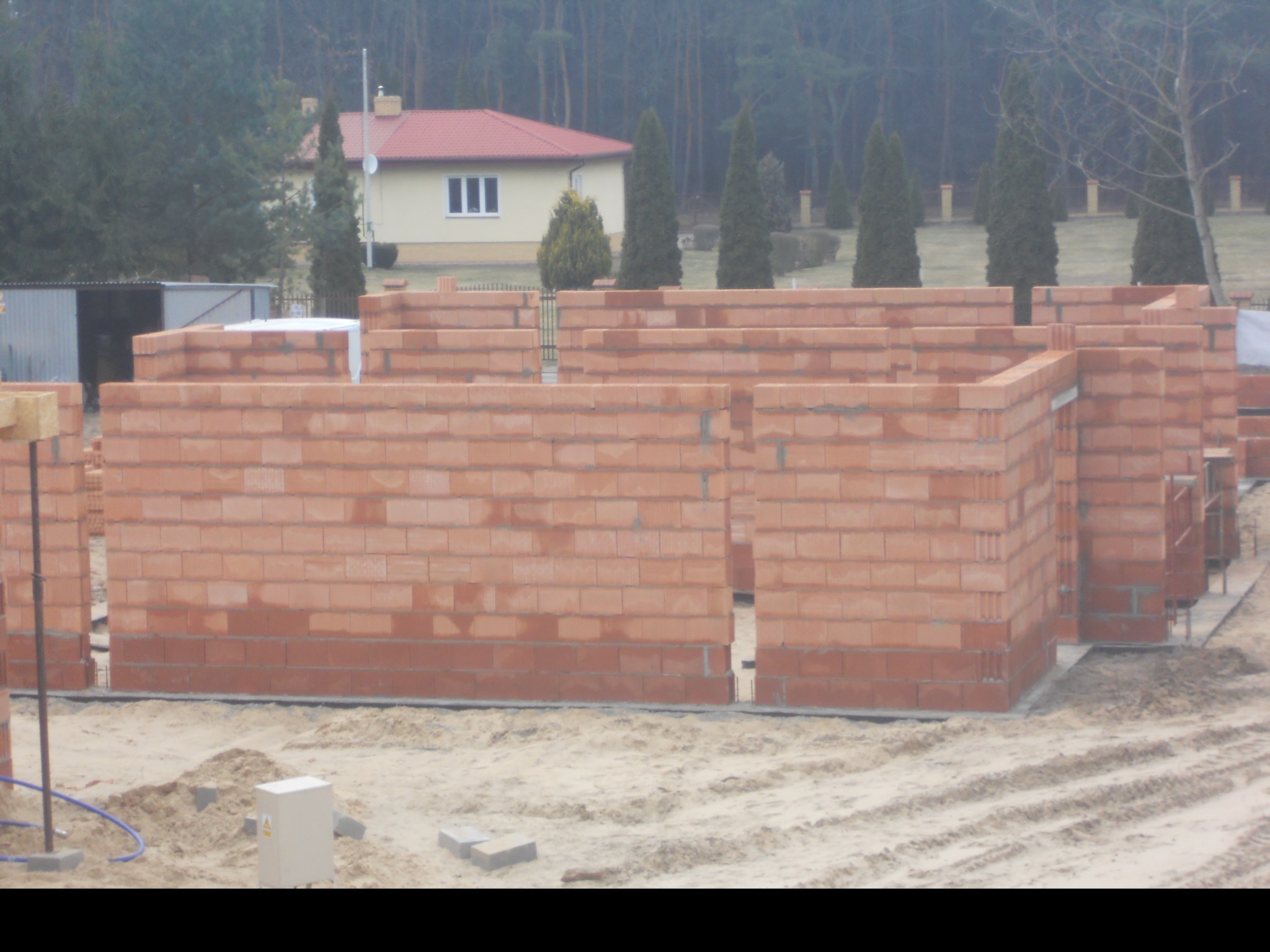 Wznoszone ściany parterowego domu z czerwonej cegły na fundamencie, widoczne zarysy pomieszczeń, w tle dom jednorodzinny z czerwonym dachem i las.