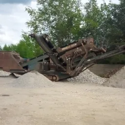 Przenośny przesiewacz Powerscreen z taśmociągiem, usypane pryzmy sortowanego kruszywa na placu, widok na zieleń drzew w tle pod pochmurnym niebem.