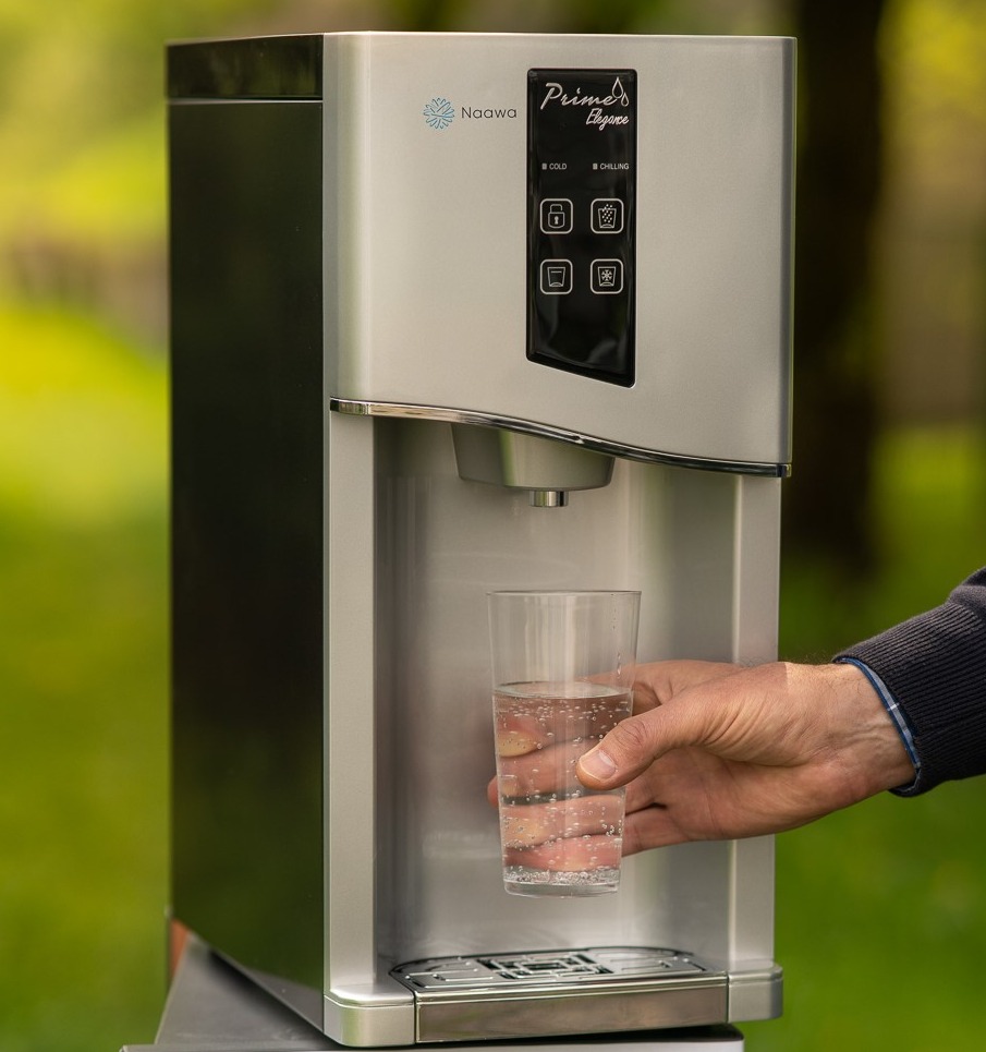 Srebrny dystrybutor wody Naowa Prime Elegance z czarnym panelem sterowania, na tle zieleni w Krzeszowicach, podczas napełniania szklanki wodą przez męską dłoń.