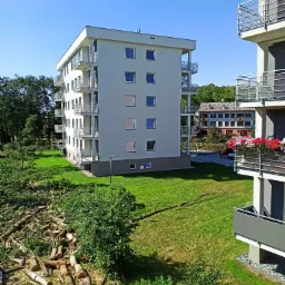 Stosy pociętych pni drzew leżą na trawniku przed nowoczesnym budynkiem mieszkalnym z balkonami, widoczne również inne budynki w tle.