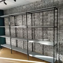 Metalowy regał sklepowy z szarymi półkami na tle ściany z szarej cegły dekoracyjnej, oświetlenie punktowe na suficie.
