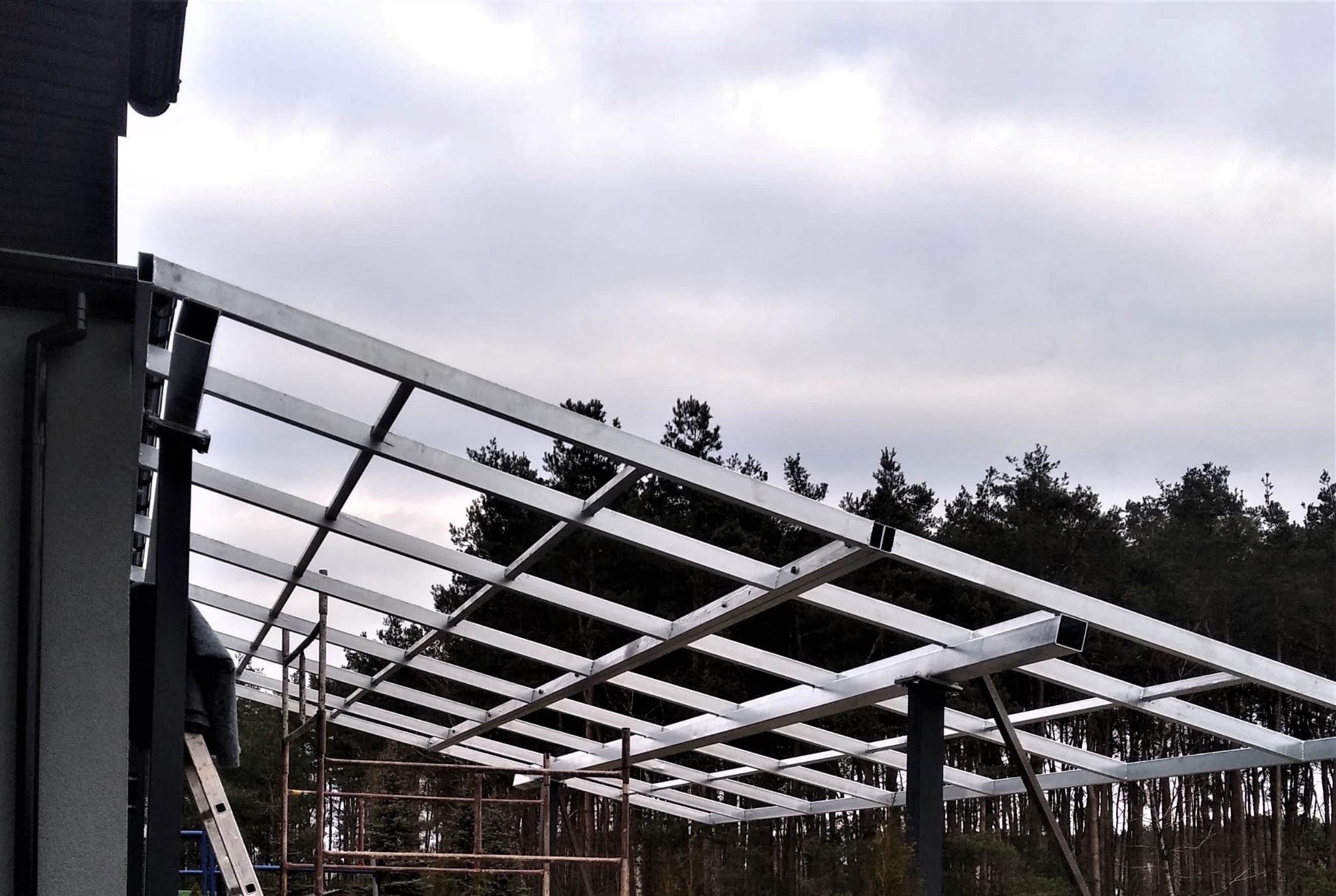 Stalowa konstrukcja pergoli w trakcie montażu, widoczne spawane profile kwadratowe, drabina i rusztowanie w tle, pochmurne niebo.