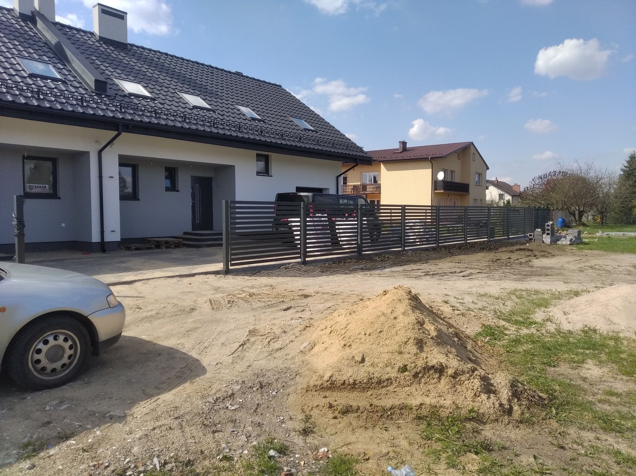 Nowoczesne ogrodzenie z poziomych metalowych profili w kolorze grafitowym, oddzielające posesję od ulicy, widoczne na tle domu z ciemną dachówką i stertą piasku.
