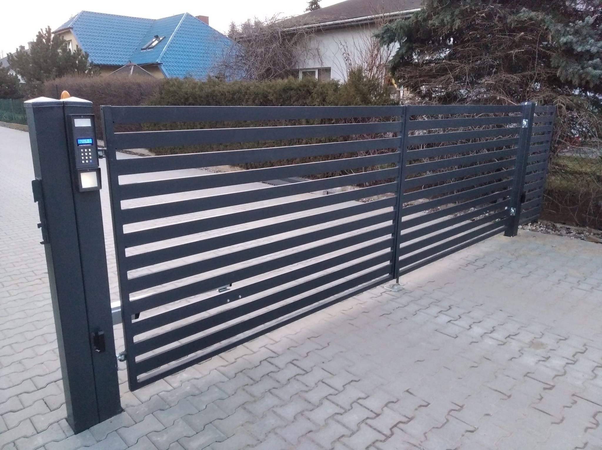 Nowoczesna brama przesuwna z poziomych profili metalowych, zintegrowana z panelem sterowania dostępem, na tle kostki brukowej i zieleni.