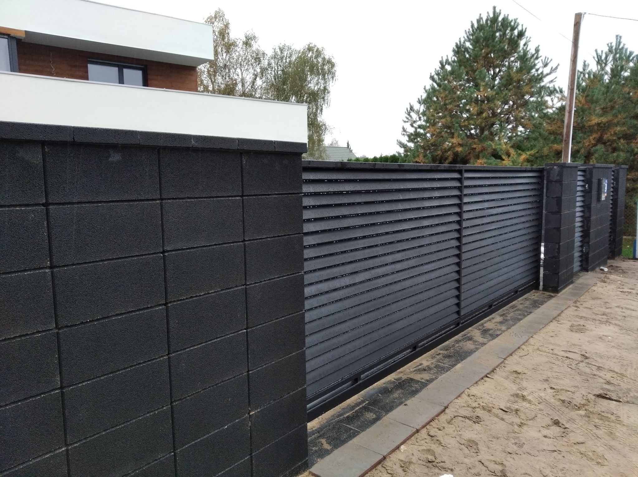 Nowoczesna, przesuwna brama w kolorze antracytowym, z poziomymi lamelami, zamontowana w Warszawie. Brama osadzona jest między słupkami z czarnych bloczków betonowych, tworzących spójny wygląd...