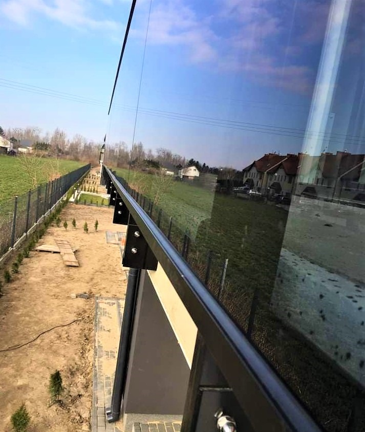 Nowoczesne, szklane ogrodzenie z czarnymi elementami mocującymi, widok z góry na posesję z trawnikiem i ogrodzeniem panelowym w tle.