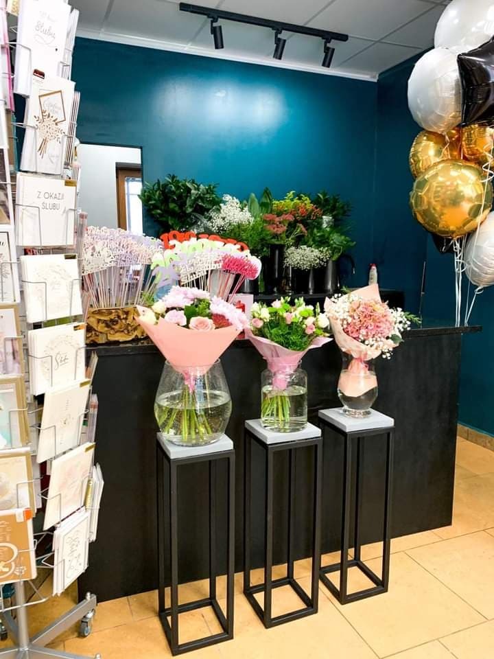 Kwiaciarnia: Trzy bukiety w szklanych wazonach na czarnych, minimalistycznych stojakach, kartki okolicznościowe na metalowym stojaku oraz balony wypełnione helem w tle, tworzące kompozycję prezentową.