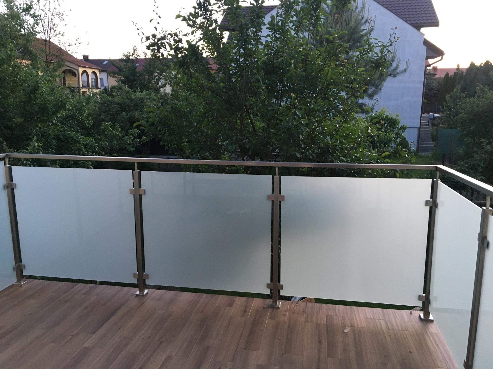 Szklana balustrada balkonowa z elementami ze stali nierdzewnej, widok z zewnątrz na zieleń i zabudowania.