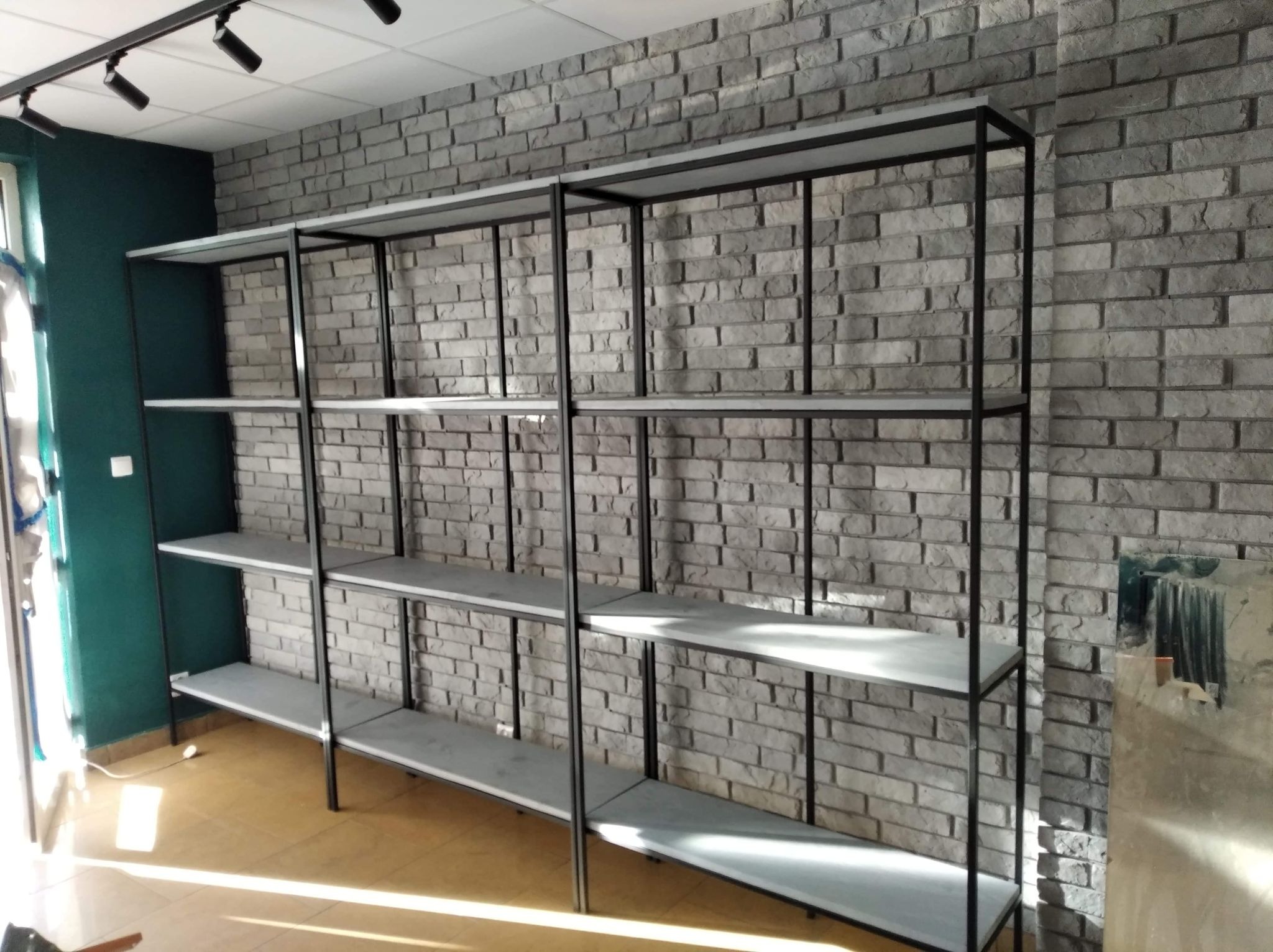 Metalowy regał sklepowy z szarymi półkami na tle ściany z szarej cegły dekoracyjnej, oświetlenie punktowe na suficie.