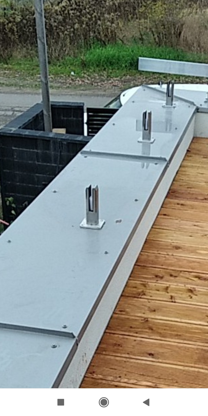 Metalowe słupki balustrady szklanej, zamontowane na szarym podwyższeniu, przylegającym do drewnianego tarasu. Widoczne śruby mocujące i fragment otoczenia z zielenią.
