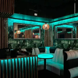 Wnętrze lokalu z drewnianymi elementami, zieloną tapetą w liście i oświetleniem LED w kolorze turkusowym, lustra w ozdobnych ramach odbijające lampy z drewnianych listewek.