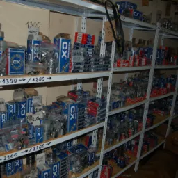 Magazyn z półkami pełnymi pudełek i opakowań z łożyskami, pasami klinowymi i kołami, w większości marki CX, z odręcznymi oznaczeniami na półkach.