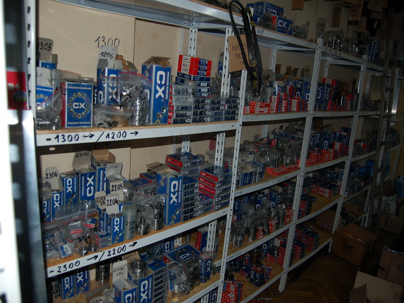 Magazyn z półkami pełnymi pudełek i opakowań z łożyskami, pasami klinowymi i kołami, w większości marki CX, z odręcznymi oznaczeniami na półkach.