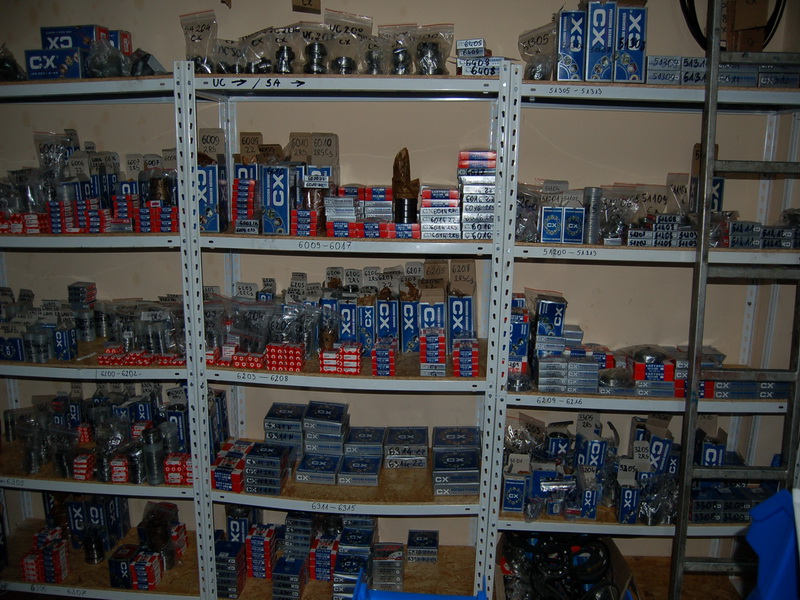 Półki regału magazynowego wypełnione zapasami łożysk, pasów i kół w opakowaniach z logo CX, ułożonych w rzędach i kolumnach, widok z perspektywy magazynu.