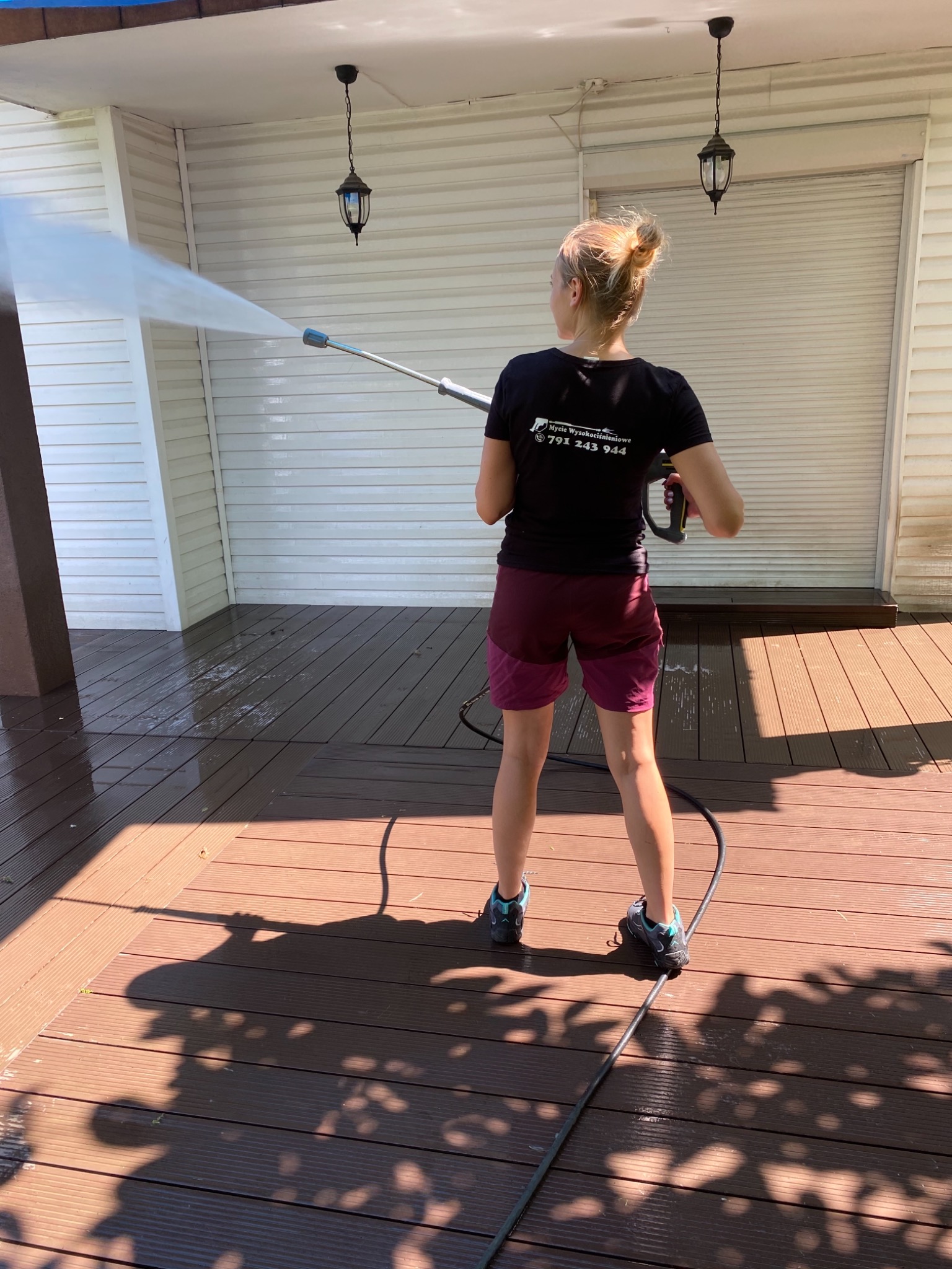 Kobieta w ciemnej koszulce z logo firmy czyści deskę tarasową za pomocą myjki ciśnieniowej, widoczne mokre deski i woda rozpryskująca się na elewacji.
