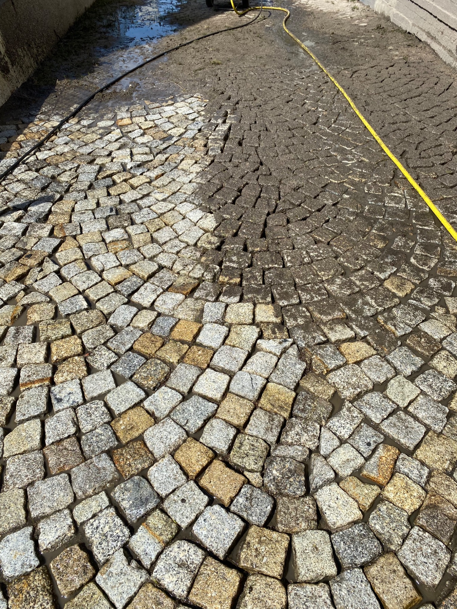 Częściowo umyta kostka brukowa z widoczną żółtą rurą i resztkami brudu w szczelinach, ukazująca kontrast przed i po czyszczeniu.