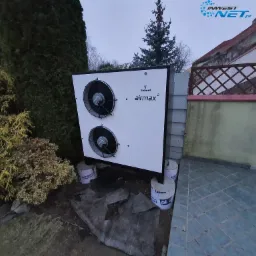 Biała pompa ciepła Galmet Airmax 2, zamontowana na betonowych wspornikach w ogrodzie, widoczna na tle krzewów i domu z ogrodzeniem z kratką.