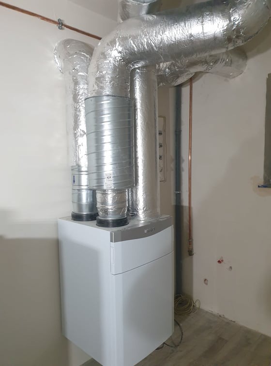 Biała jednostka rekuperacyjna Vaillant z podłączonymi, zaizolowanymi przewodami wentylacyjnymi w pomieszczeniu o surowym wykończeniu.