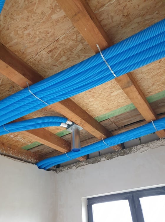 Niebieskie, karbowane rury wentylacyjne zamontowane pod drewnianym stropem, przymocowane metalowymi opaskami, z widocznym elementem łączącym rury w jedną, pionową rurę spiro.