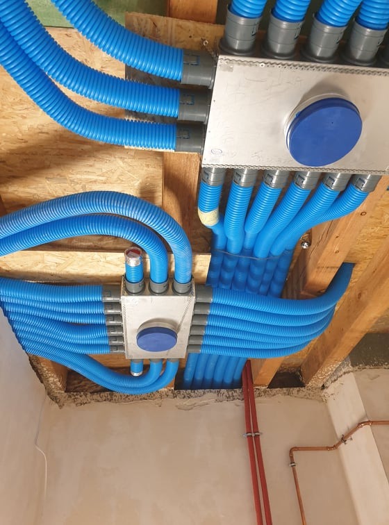 Widok instalacji rekuperacji z licznymi niebieskimi przewodami karbowanymi, rozchodzącymi się od centralnych skrzynek rozdzielczych, zamontowanych do drewnianego stropu, widoczne również rury...