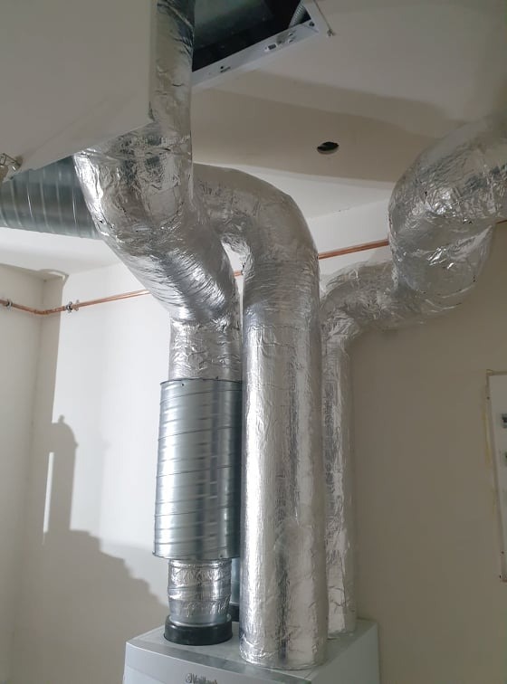System rur wentylacyjnych i rekuperacyjnych, owiniętych srebrną folią izolacyjną, podłączonych do urządzenia Vaillant, z widoczną miedzianą rurką na ścianie.