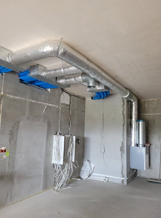 Niedokończona instalacja systemu wentylacyjnego z rekuperacją: srebrne rury izolowane termicznie pod sufitem, niebieskie elementy mocujące, rekuperator na ścianie, surowe ściany i kable elektryczne.