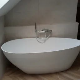 Wolnostojąca, owalna wanna z chromowaną armaturą i słuchawką prysznicową na tle beżowych płytek ściennych i podłogi z paneli ułożonych w jodełkę.