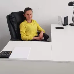Kobieta w żółtej koszuli siedzi na czarnym skórzanym krześle za białym biurkiem z kalkulatorem i czystą kartką papieru, lampa biurkowa w tle.