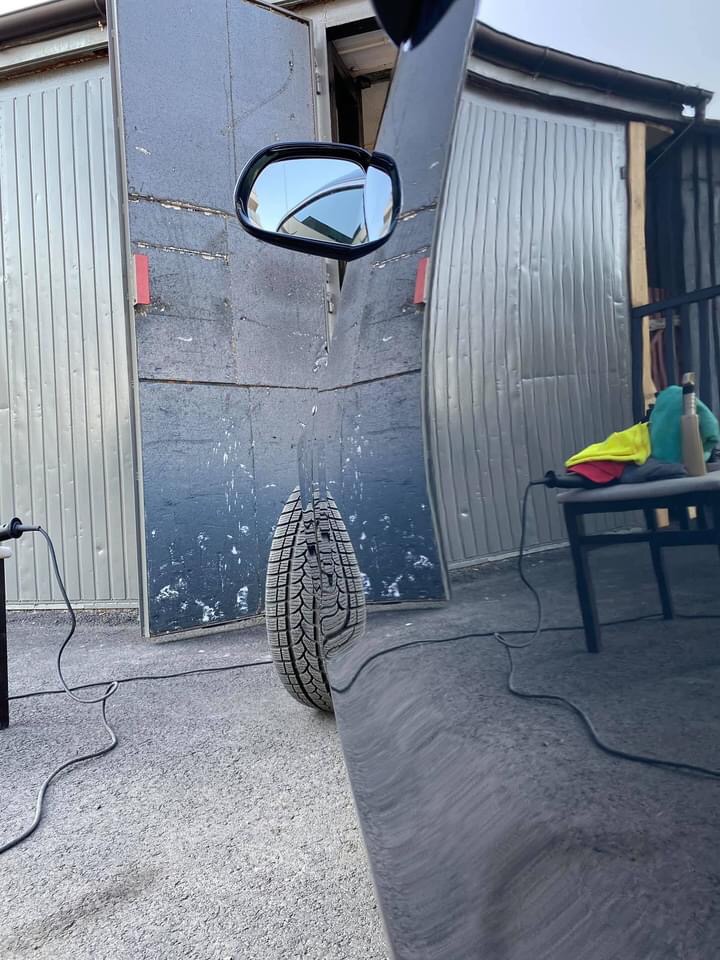 Błyszczący bok czarnego samochodu po polerowaniu, odbijający oponę, garaż blaszany i niebieskie niebo w lusterku bocznym.