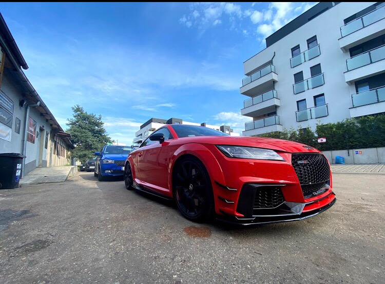 Czerwone Audi TT RS zaparkowane na zewnątrz, z widocznymi detalami karoserii i czarnymi felgami, na tle budynku mieszkalnego i innych samochodów.