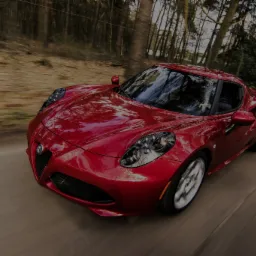 Czerwone sportowe auto Alfa Romeo 4C w ruchu na krętej drodze, widok z przodu, rozmyte tło drzew.