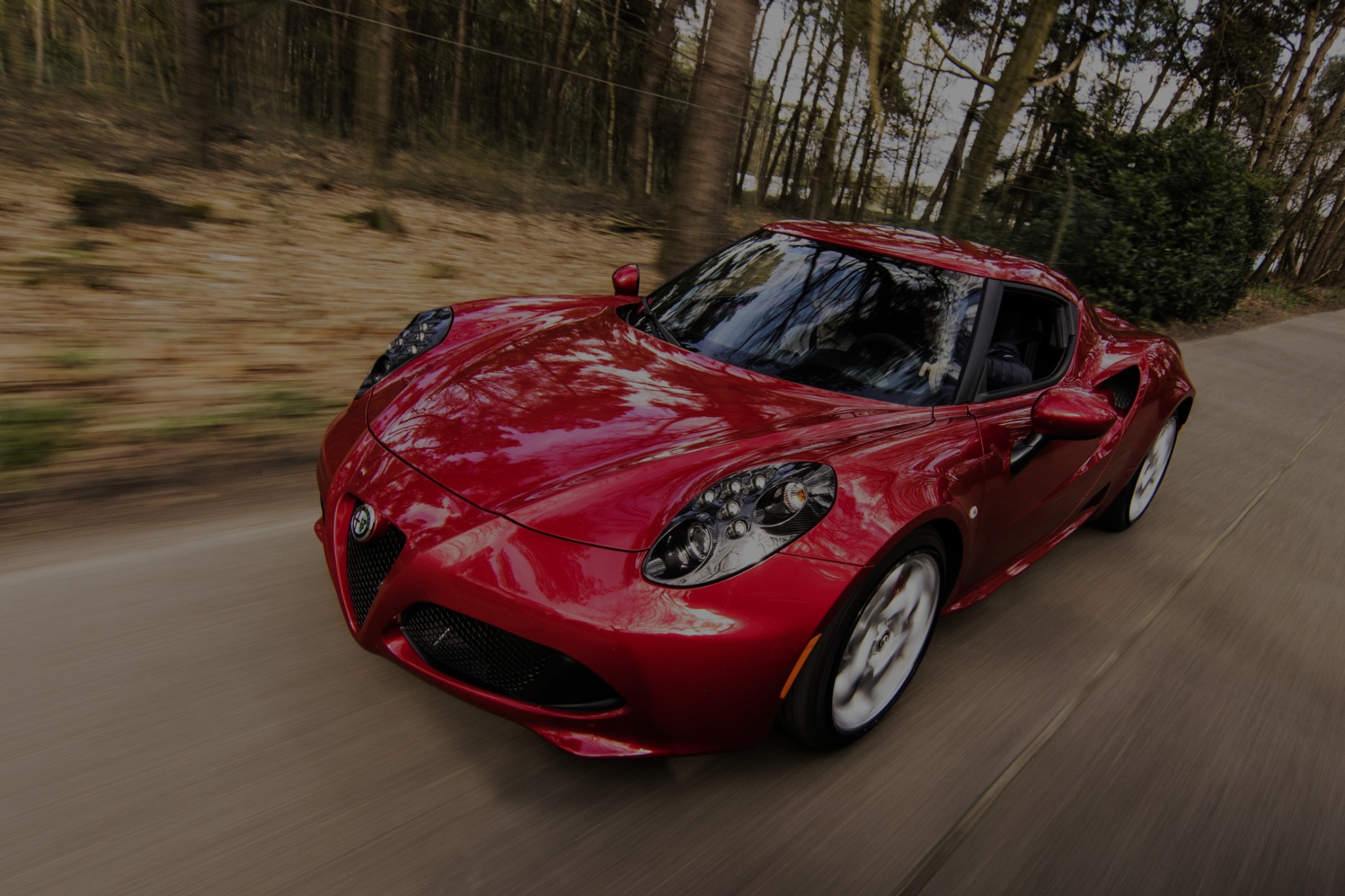 Czerwone sportowe auto Alfa Romeo 4C w ruchu na krętej drodze, widok z przodu, rozmyte tło drzew.