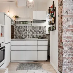 Biała kuchnia z wzorzystą ścianą nad blatem, ceglanym filarem i balkonem w tle, oświetlona wiszącą lampą drucianą.