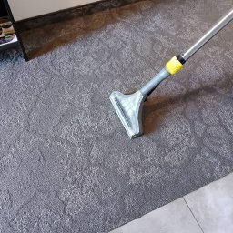 TIP TOP CLEANING SERVICES AGNIESZKA ROGAL-MOŃ - Szczegółowe ujęcie czyszczenia szarego dywanu odkurzaczem piorącym z żółtymi elementami, ukazujące profesjonalny sprzęt w trakcie pracy.