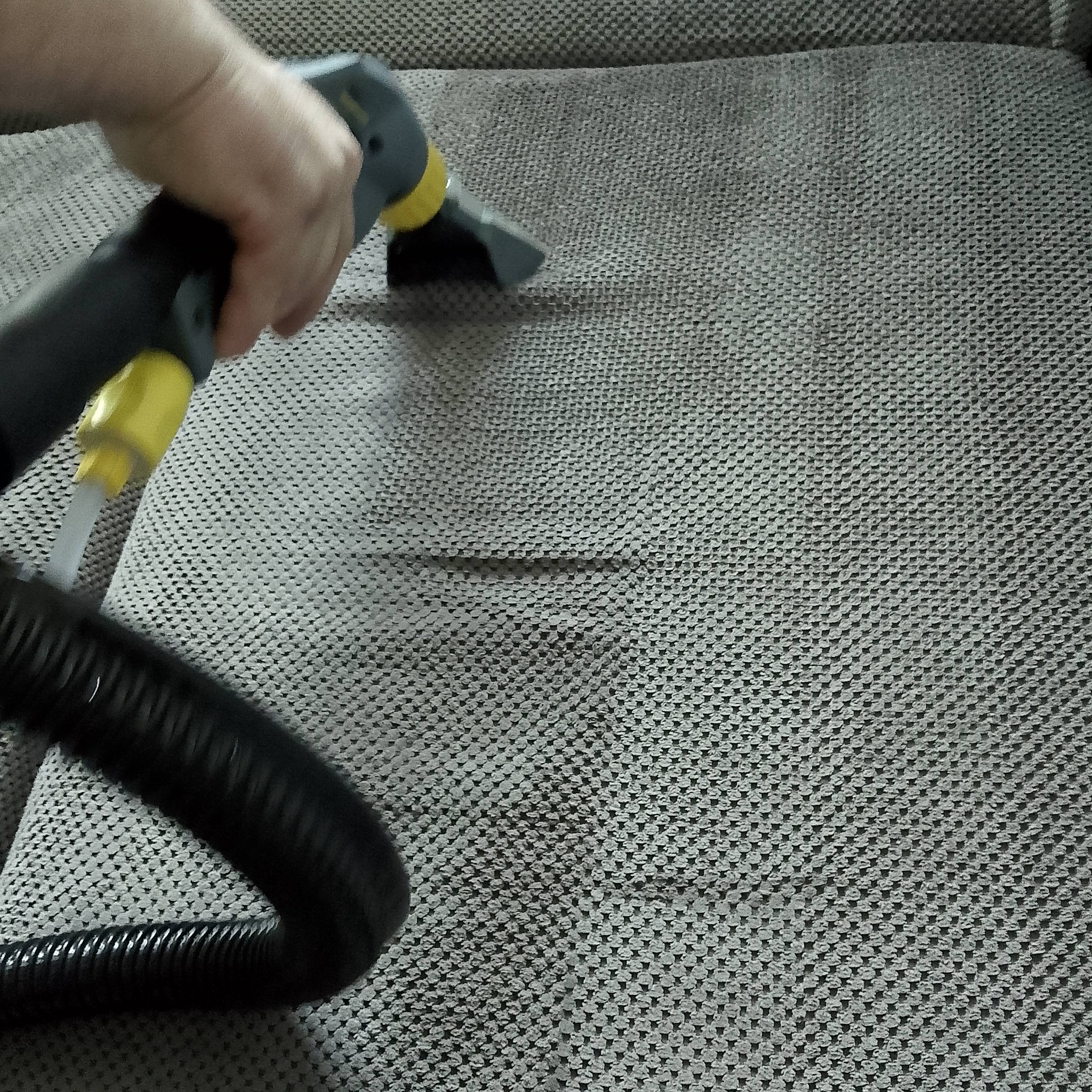 Czyszczenie sofy z szarej tkaniny odkurzaczem piorącym. Widoczny wąż, dysza i ręka osoby obsługującej urządzenie.