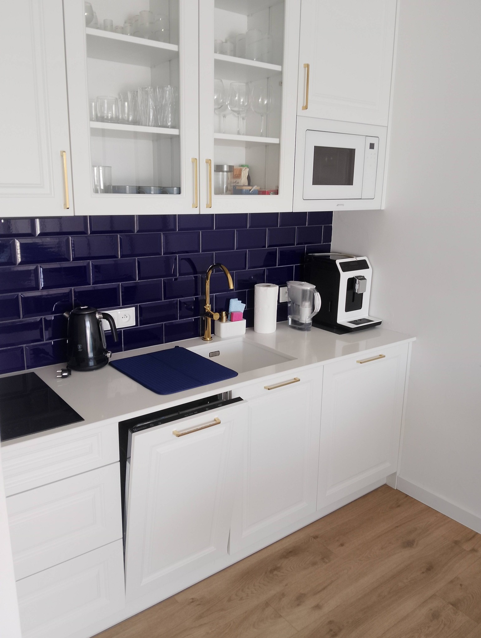 Nowoczesna, biała kuchnia z granatowymi płytkami i złotymi akcentami. Widoczny ekspres do kawy, czajnik i zlew z ociekaczem. Otwarta zmywarka.