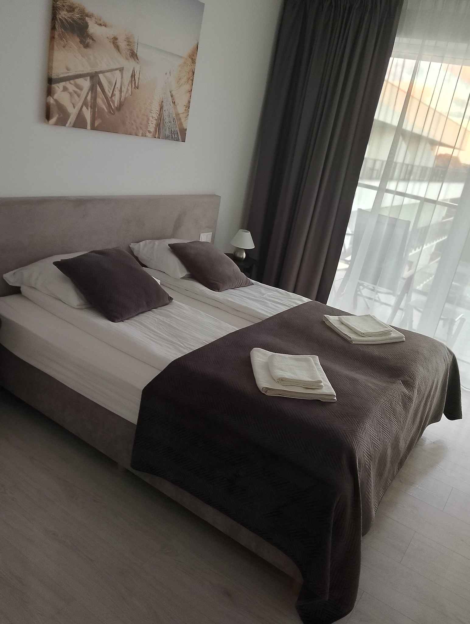 Ustawione łóżko w pokoju hotelowym z brązową narzutą i ręcznikami, widok na balkon przez zasłony. Na ścianie obraz z motywem plaży.