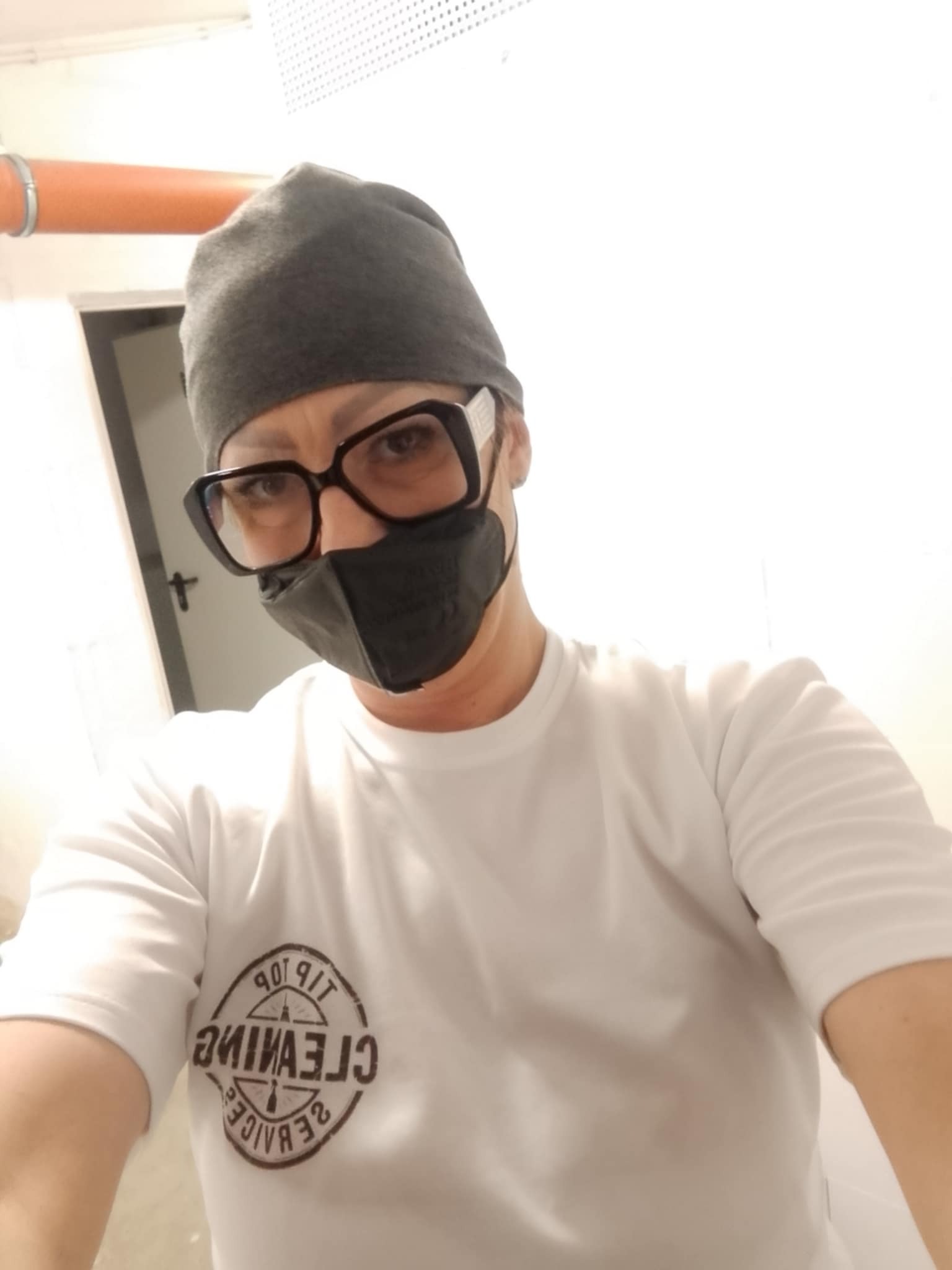 Osoba w czapce i masce ochronnej, z logo firmy sprzątającej na białej koszulce, patrzy w obiektyw. Widoczny fragment rury w tle.