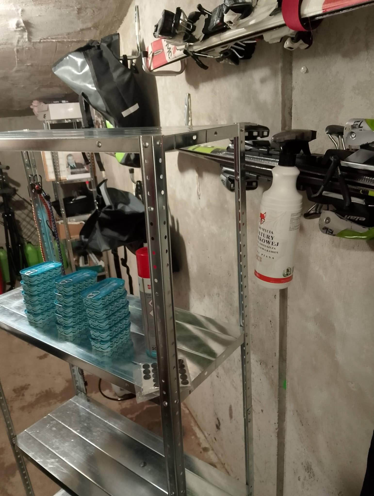 Metalowy regał w piwnicy z zapasami i sprzętem narciarskim. Widoczny środek czyszczący i puszki z farbą. Ściana betonowa, surowa. Sprzęt sportowy w tle.