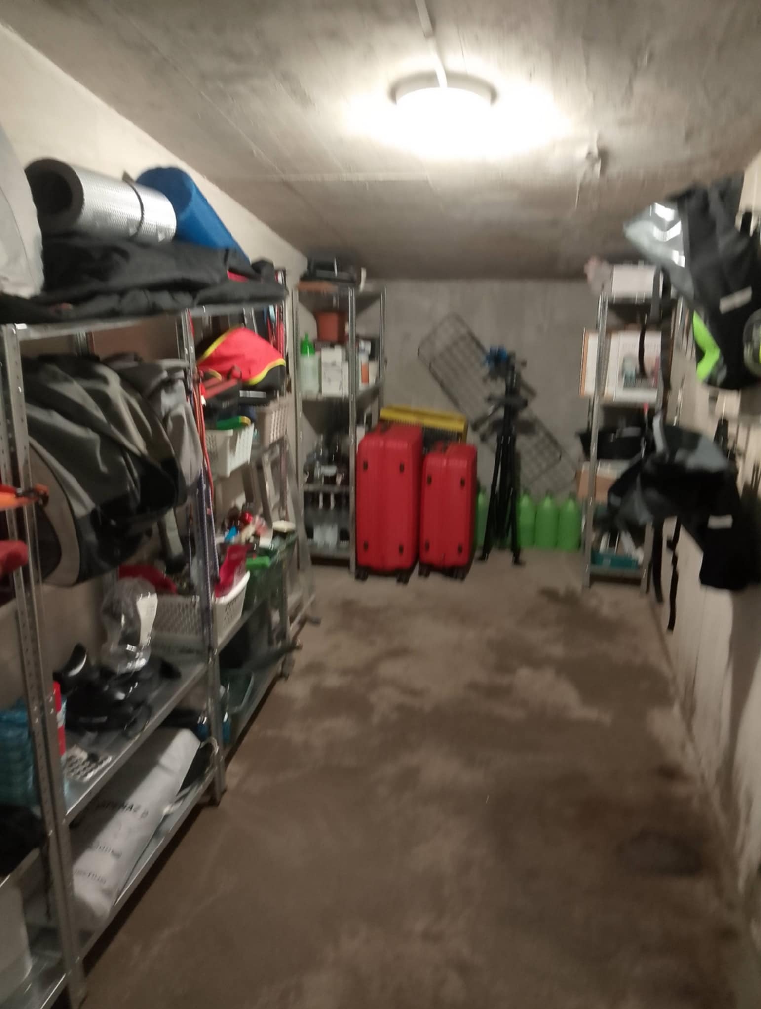 Zastawiony korytarz piwniczny z metalowymi regałami, walizkami i sprzętem. Szare ściany, sufit z lampą. Widok na zagracone pomieszczenie.