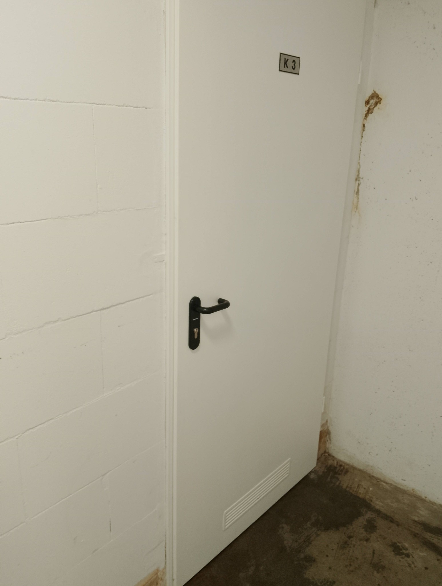 Białe drzwi z czarną klamką i oznaczeniem 'K 3' w rogu pomieszczenia. Na ścianach widoczne ślady wilgoci i zabrudzenia. Podłoga betonowa.