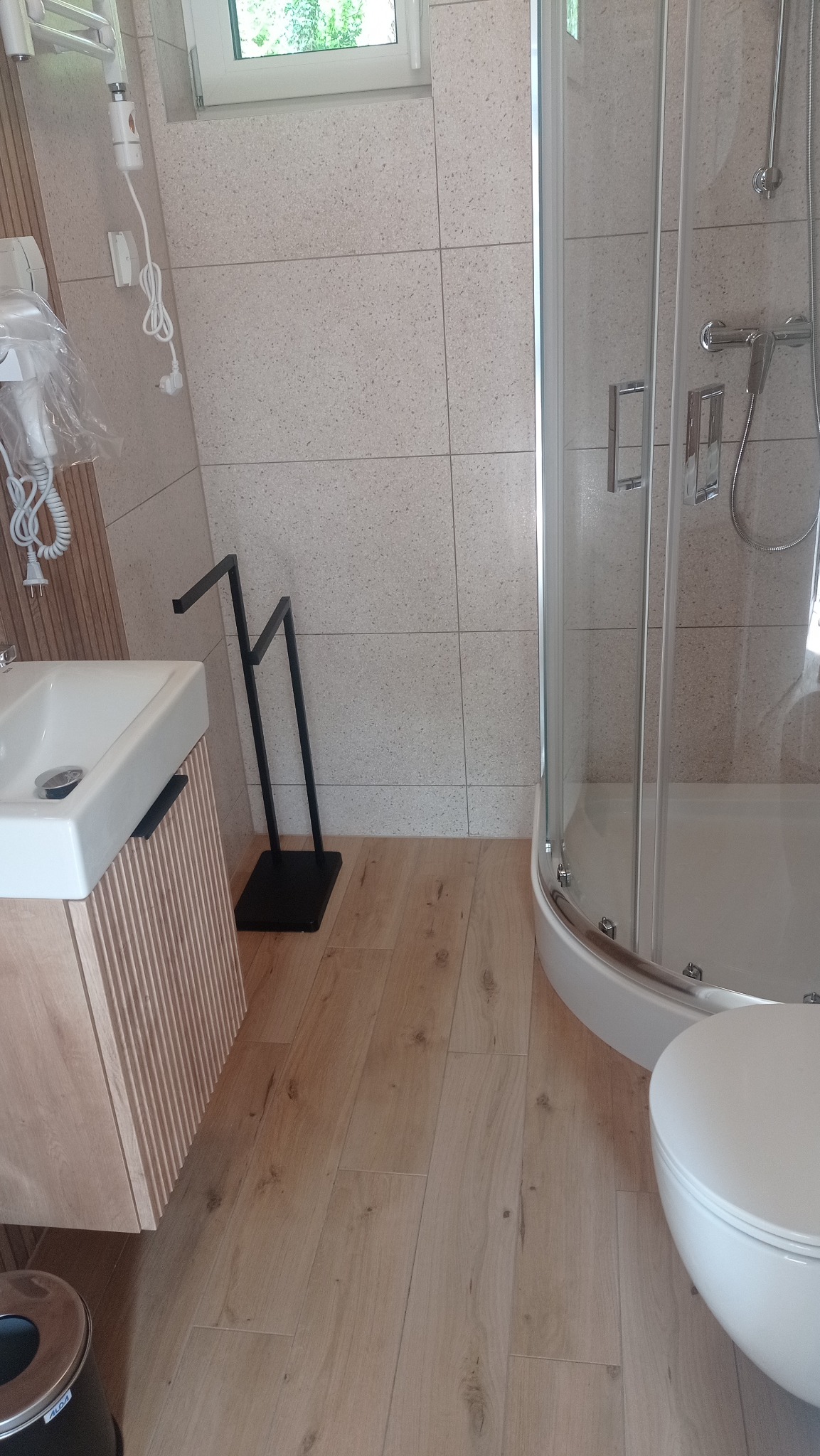 Nowoczesna łazienka z kabiną prysznicową, umywalką z szafką z lamelami i drewnopodobną podłogą. Minimalistyczny czarny wieszak na ręczniki i okno z widokiem na zieleń.