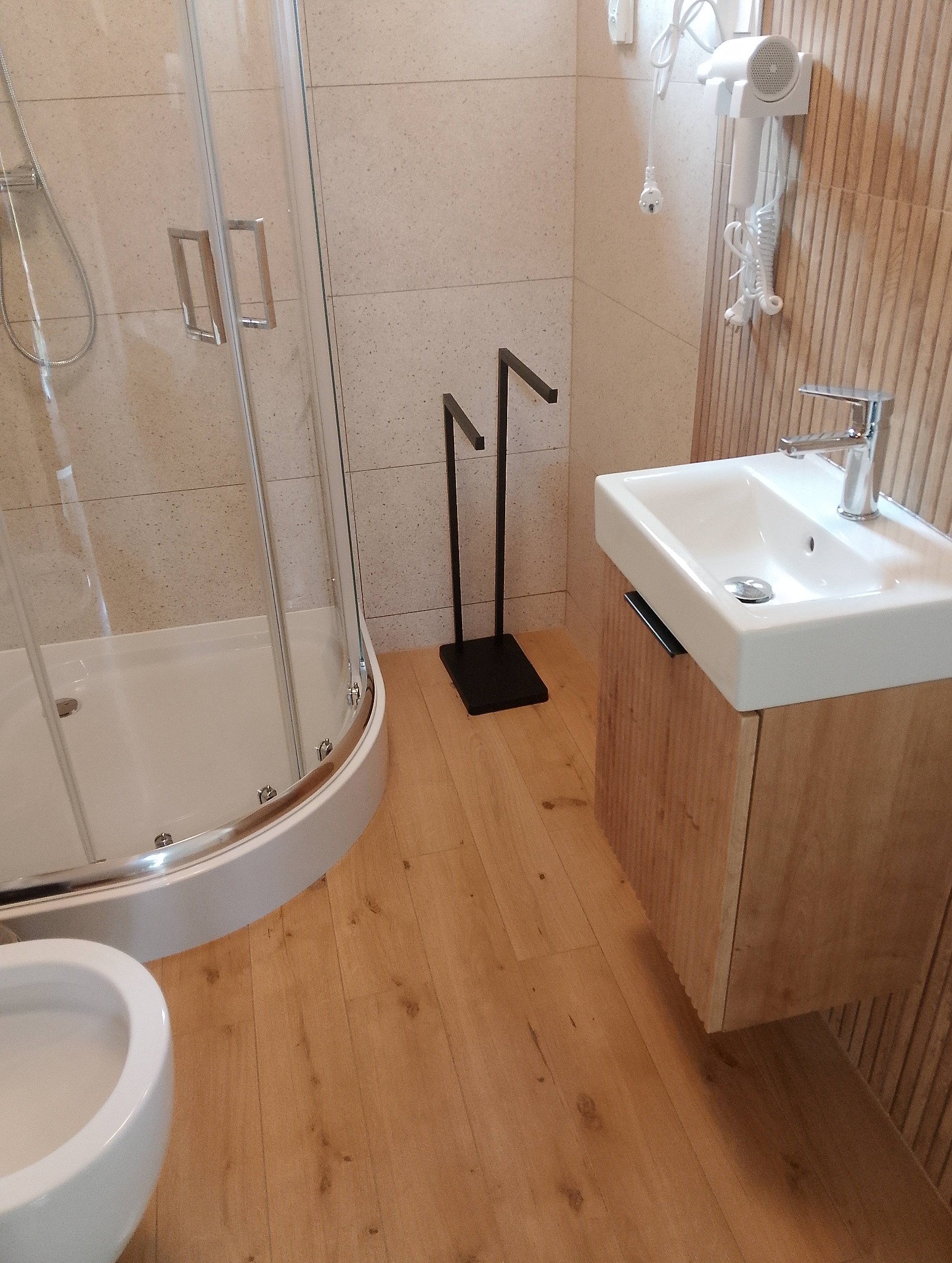 Jasna łazienka z kabiną prysznicową, umywalką na szafce z drewnianymi lamelami i suszarką do włosów na ścianie. Ujęcie z niskiego kąta.