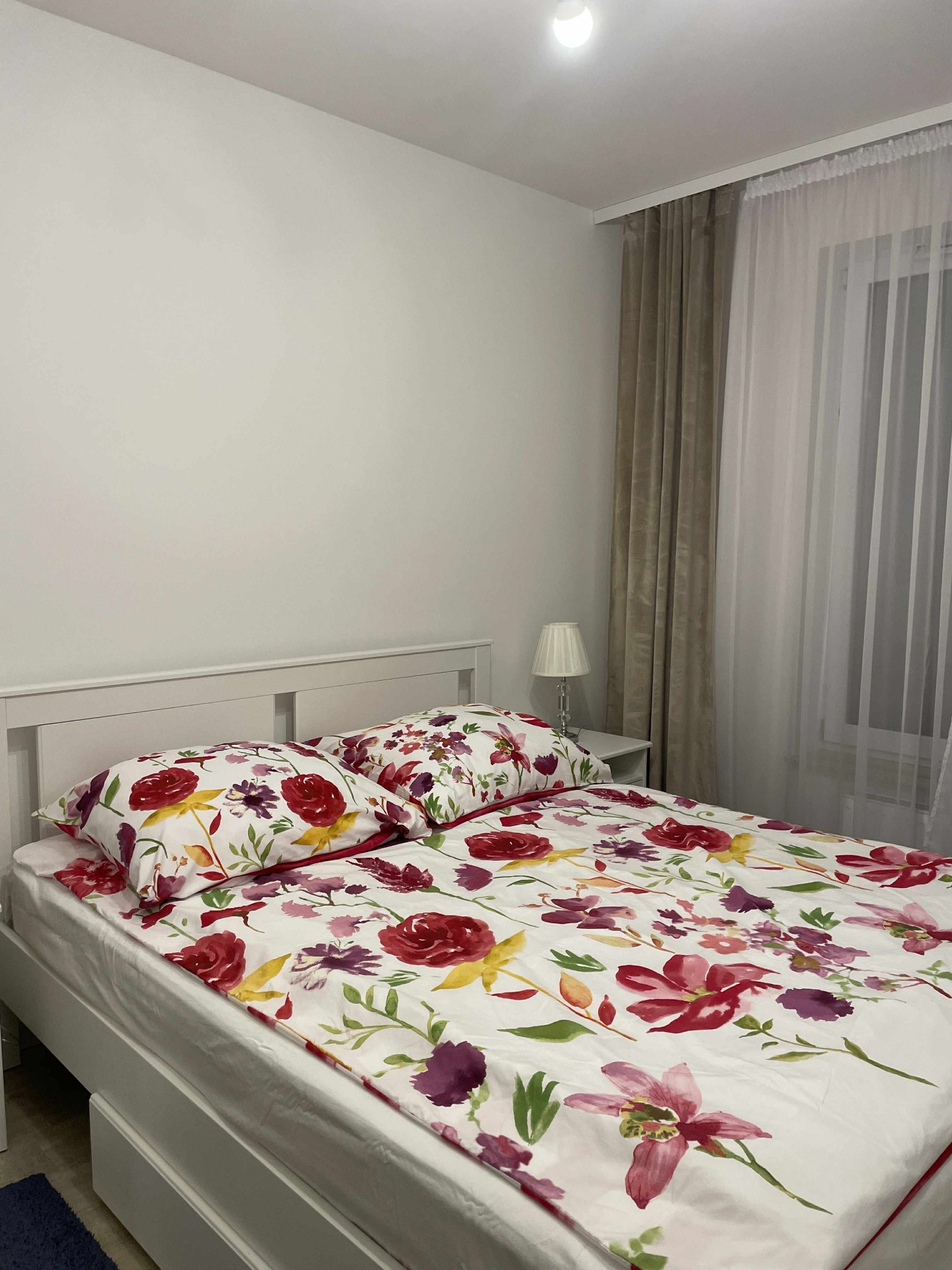 Białe łóżko z kwiatową pościelą w sypialni z beżowymi zasłonami i lampką nocną. Widok z boku, oświetlenie górne.