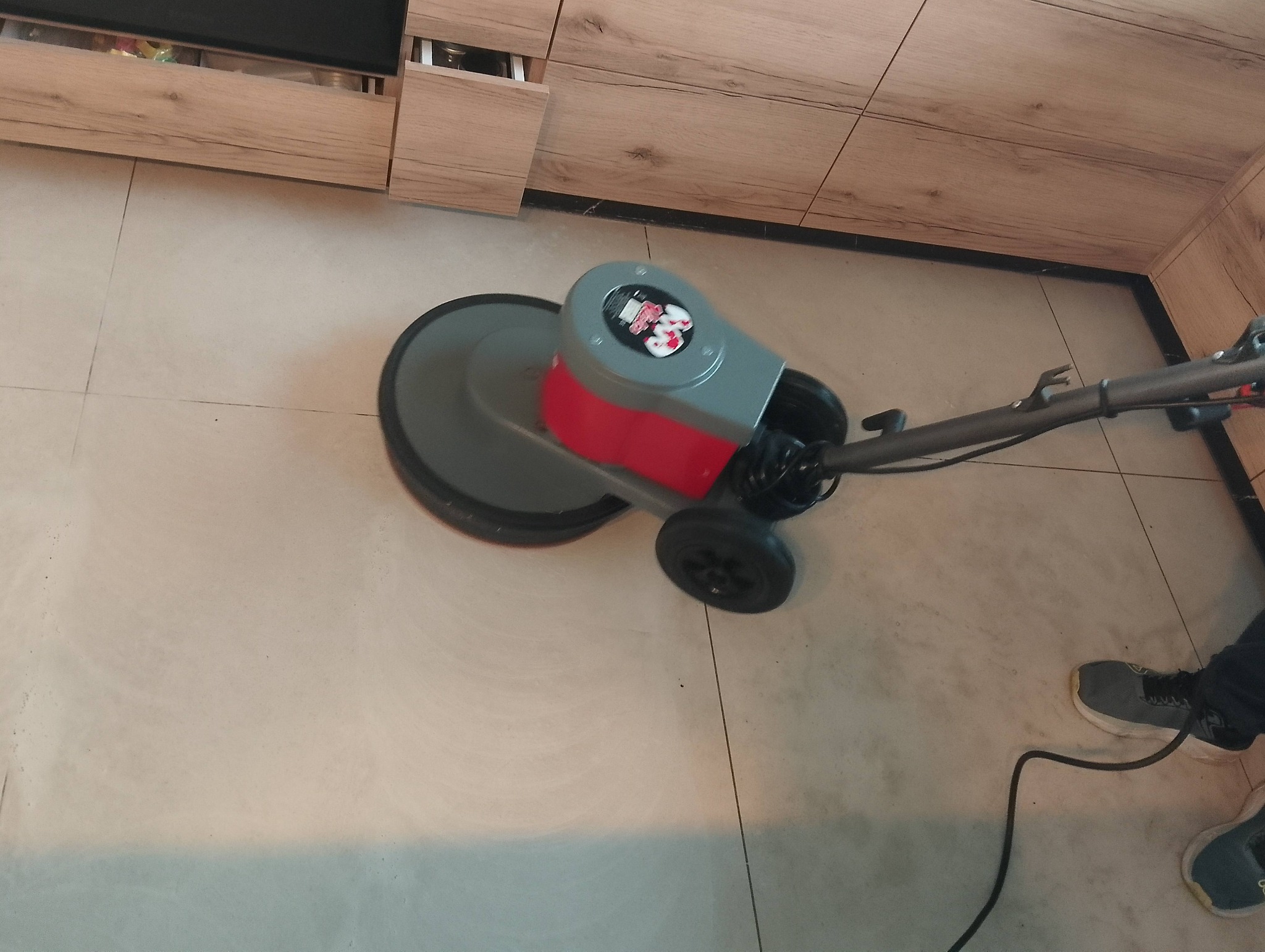 Maszyna do czyszczenia podłóg w trakcie pracy na jasnych płytkach, widoczne fragmenty szafek kuchennych i stopy osoby obsługującej urządzenie.