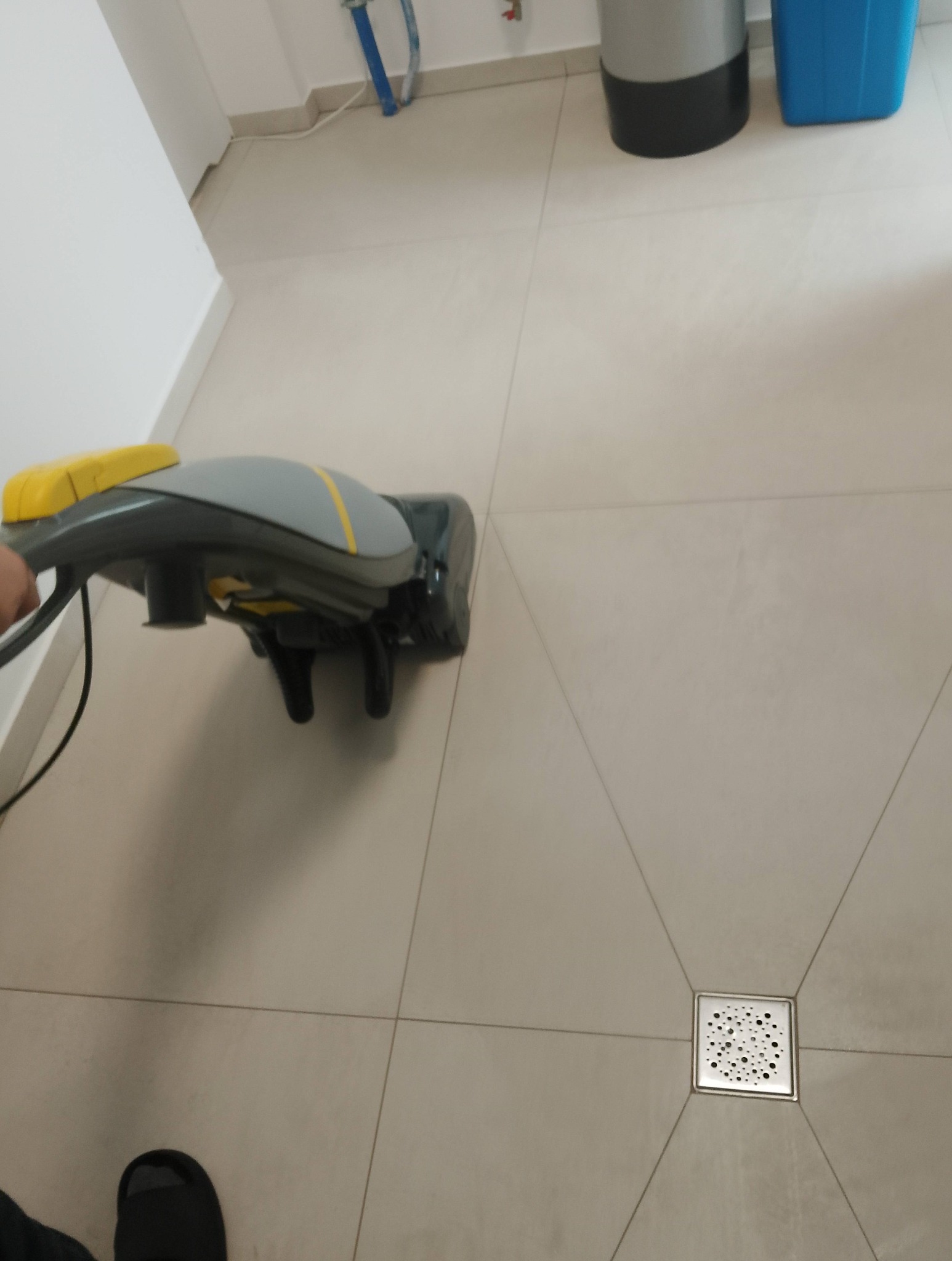 Maszyna do czyszczenia podłóg w akcji na jasnych, kwadratowych płytkach. Widoczny odpływ podłogowy i fragment nogi operatora. Sprzęt do sprzątania w tle.