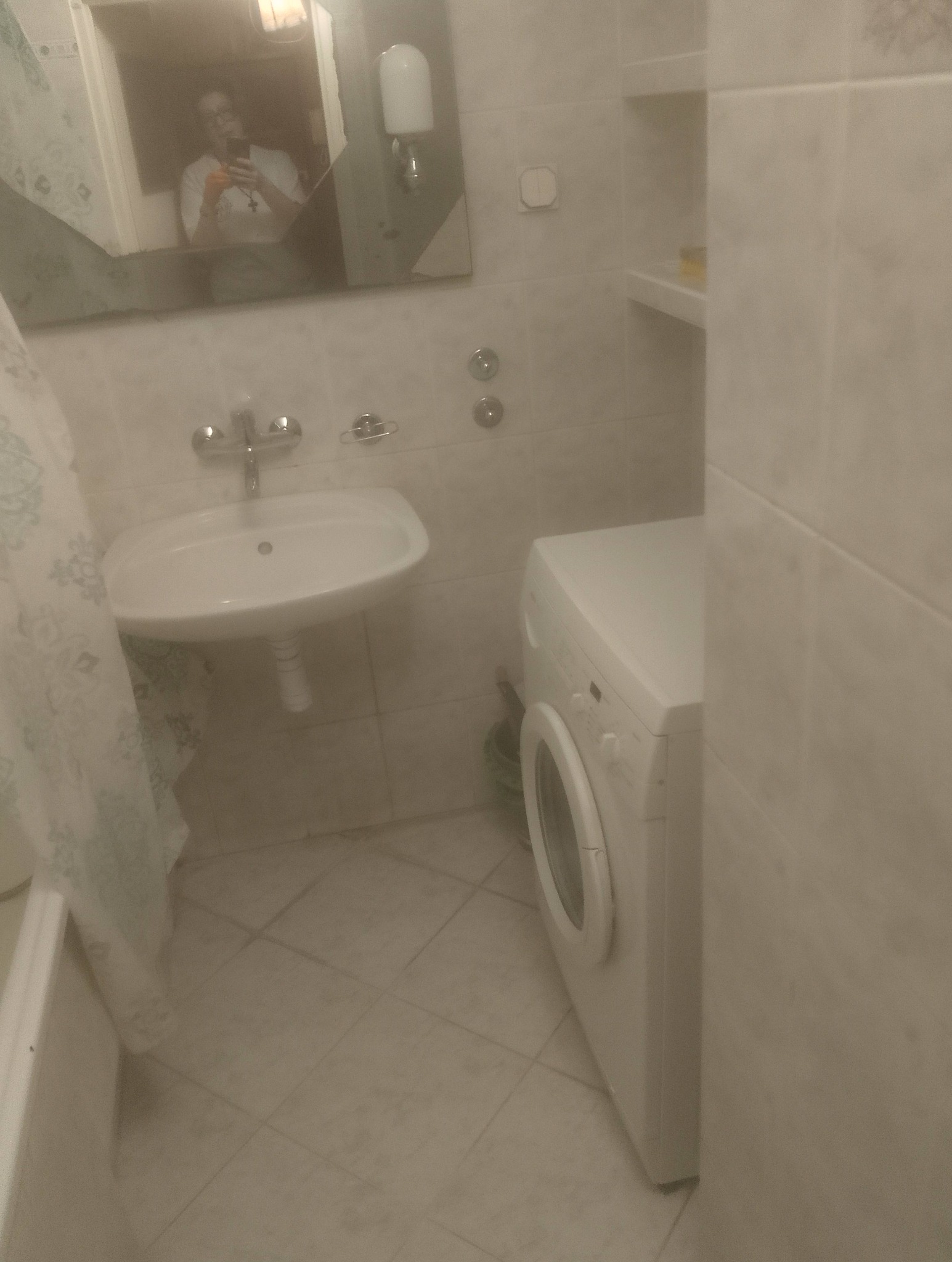 Jasna łazienka z umywalką, pralką i odbiciem osoby w lustrze. Widoczne białe płytki i elementy wyposażenia. Ujęcie z perspektywy osoby stojącej w pomieszczeniu.