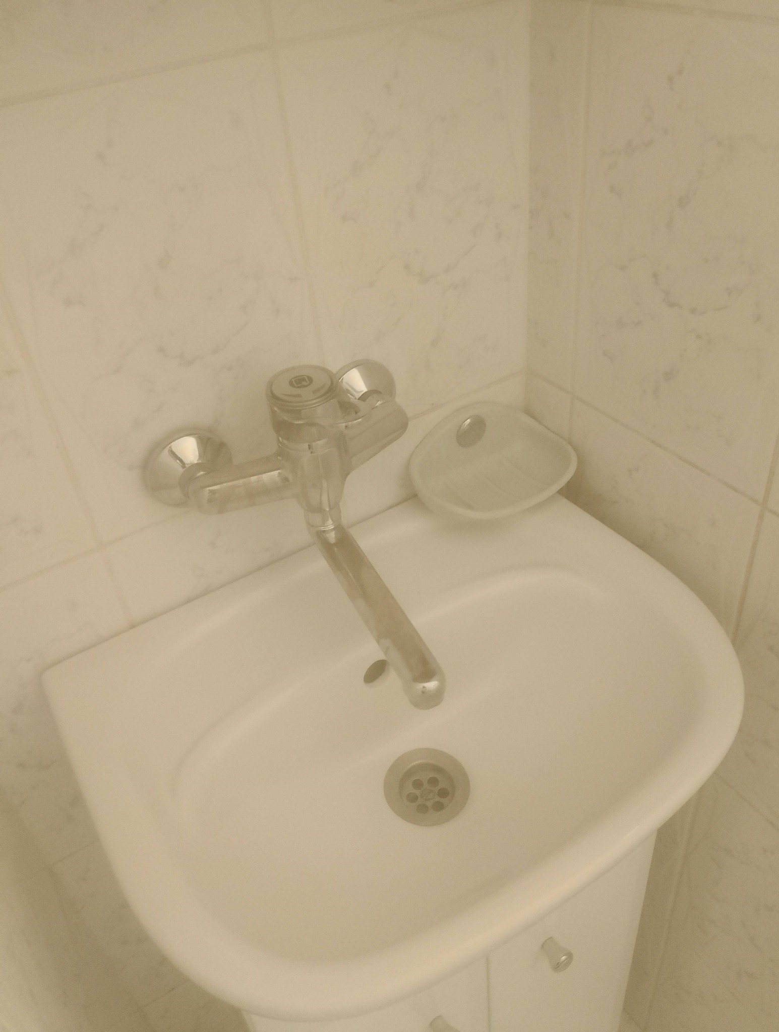 Biała umywalka z chromowaną baterią i mydelniczką na tle marmurkowych płytek. Ujęcie z góry, widoczny odpływ i detale armatury.