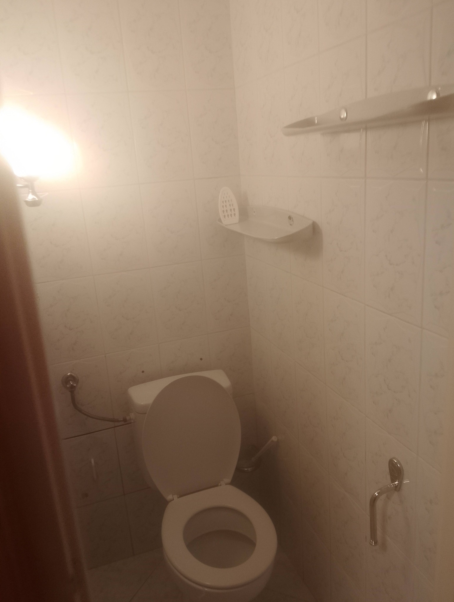 Toaleta z otwartą klapą w łazience wyłożonej jasnymi, kwadratowymi płytkami. Nad toaletą półka z akcesoriami. Oświetlenie górne. Ujęcie z dołu.