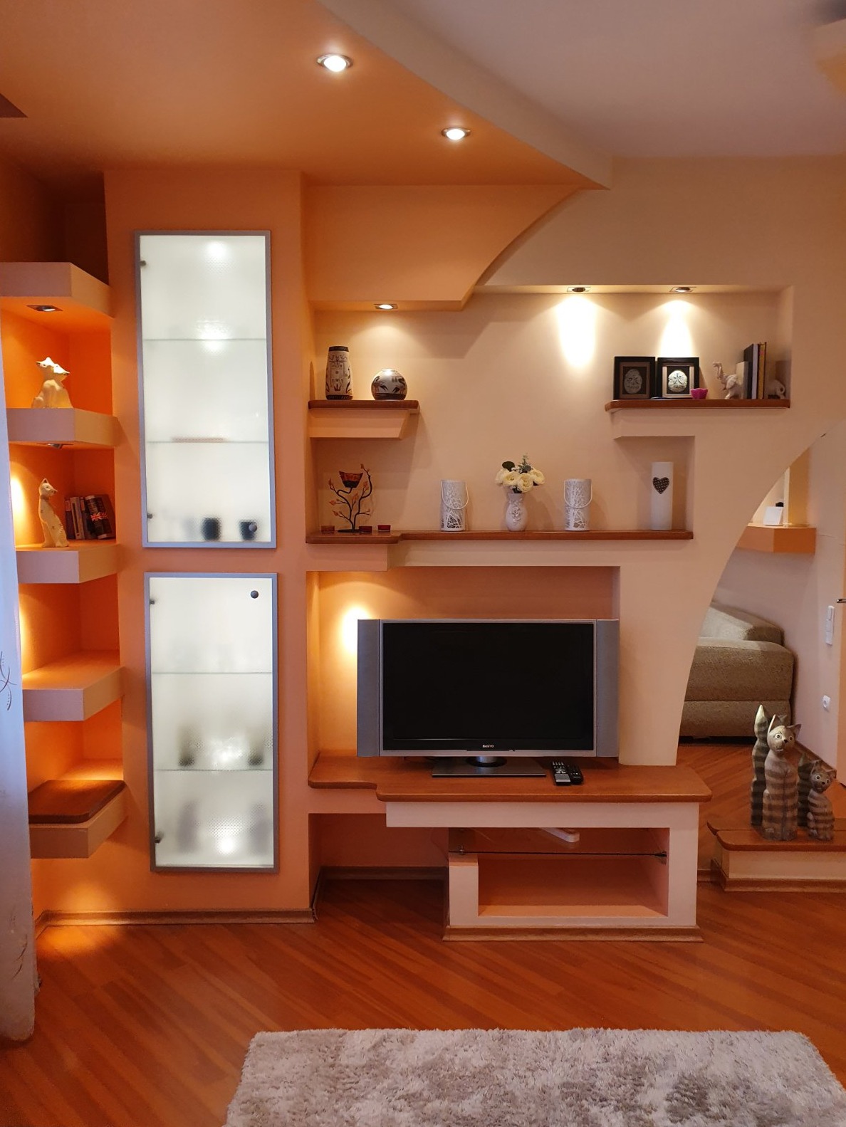 Jasne wnętrze salonu z telewizorem, podświetlanymi półkami dekoracyjnymi z bibelotami i szklanymi witrynami, oświetlenie punktowe i drewniana podłoga.